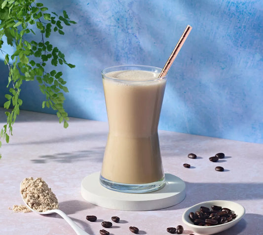 Glass of Herbalife Nutrition Formula 1 Café Latte shake mix with a straw, surrounded by coffee beans and a spoonful of powder on a light blue background.