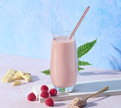 Glass of pink Herbalife Formula 1 Raspberry & White Chocolate Shake Mix smoothie with a straw, surrounded by raspberries and white chocolate pieces on a light blue background.