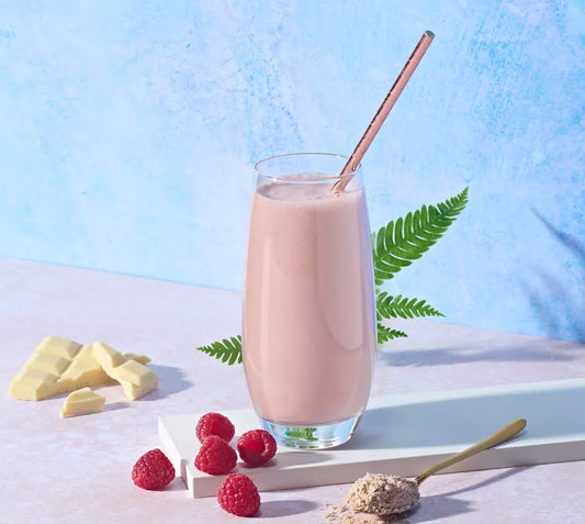 Glass of pink Herbalife Formula 1 Raspberry & White Chocolate Shake Mix smoothie with a straw, surrounded by raspberries and white chocolate pieces on a light blue background.