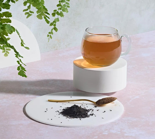 Glass of Herbalife Instant Herbal Beverage Original Flavour Large 102g Green Tea, Orange Pekoe Black Tea, Malva Flower, Hibiscous Flower and Cardomom seed beverage on a white coaster with a small amount of tea leaves and a spoon, against a light background with greenery.