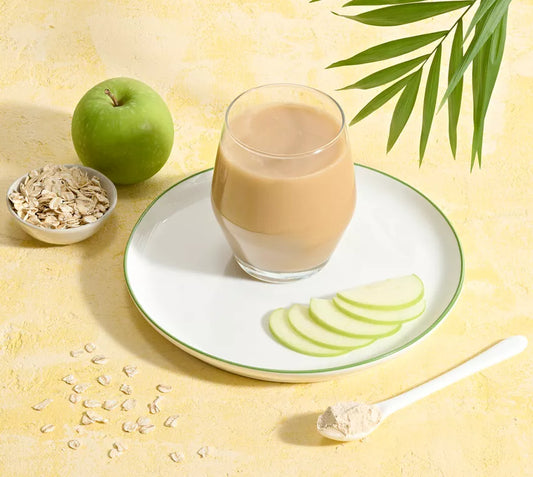 Glass of a Herbalife Oat Apple Fibre on a plate with sliced apples, an apple, and a bowl of oats on a yellow background.