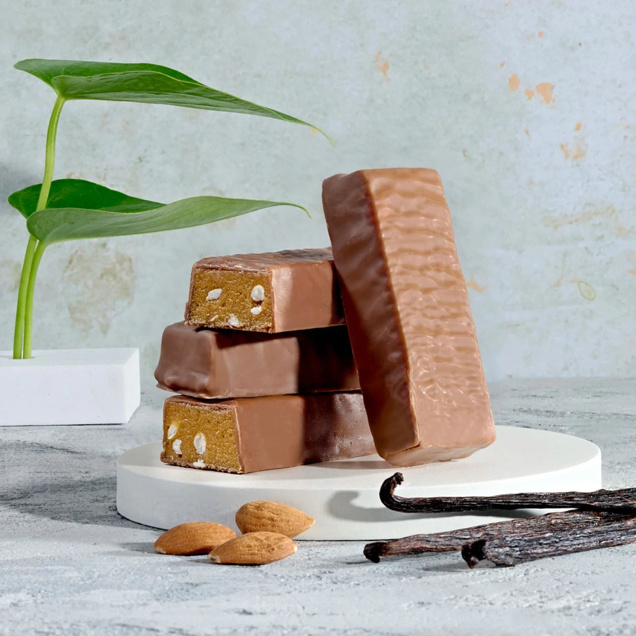 Stack of Herbalife Vanilla Almond protein bars on a white stand with vanilla beans and almonds on a light grey surface.